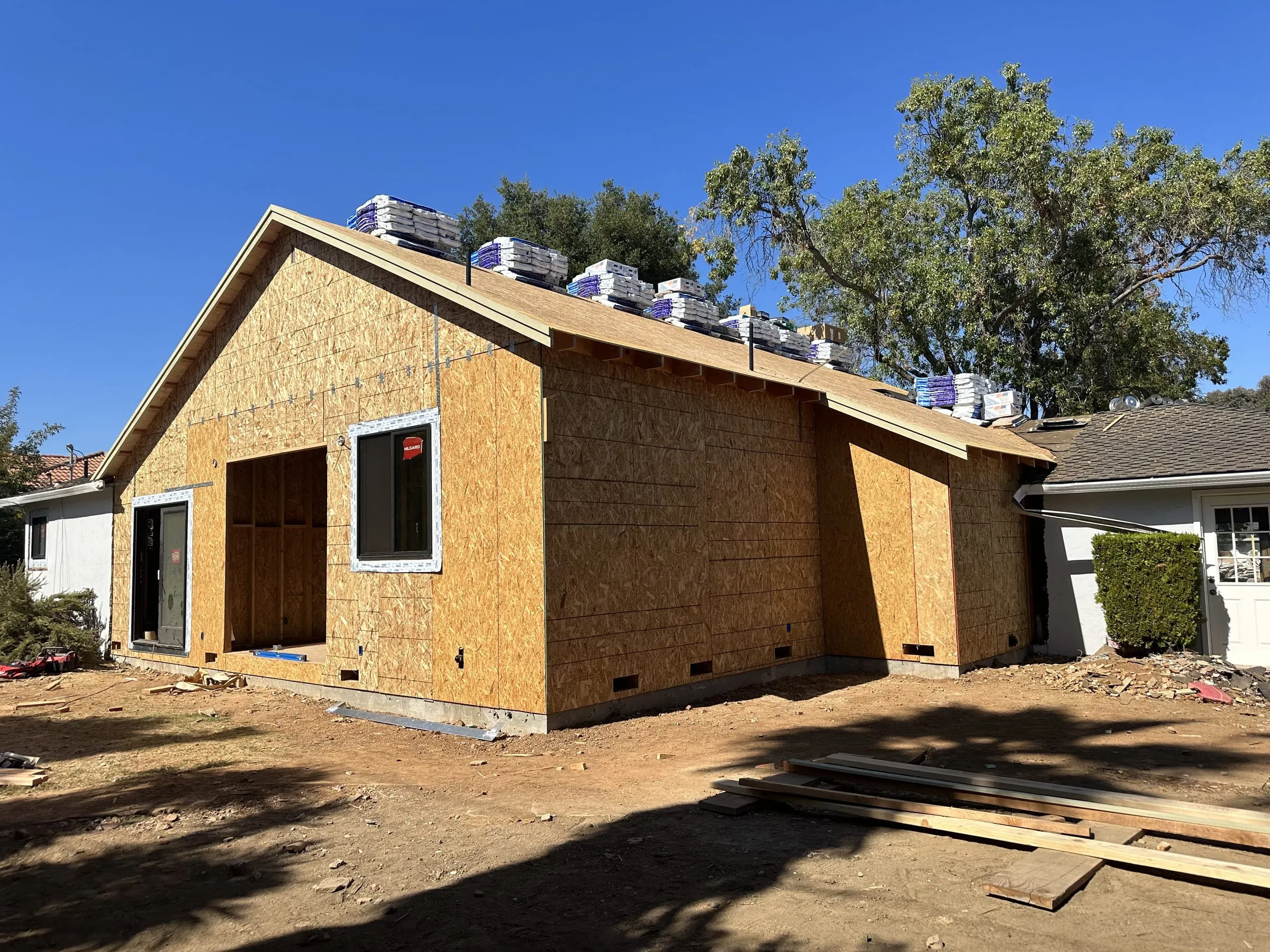 A Legacy4Builders addition framed, sheathed and dried in, tied into the existing stucco house, with shingle bundles stacked on the new roof.