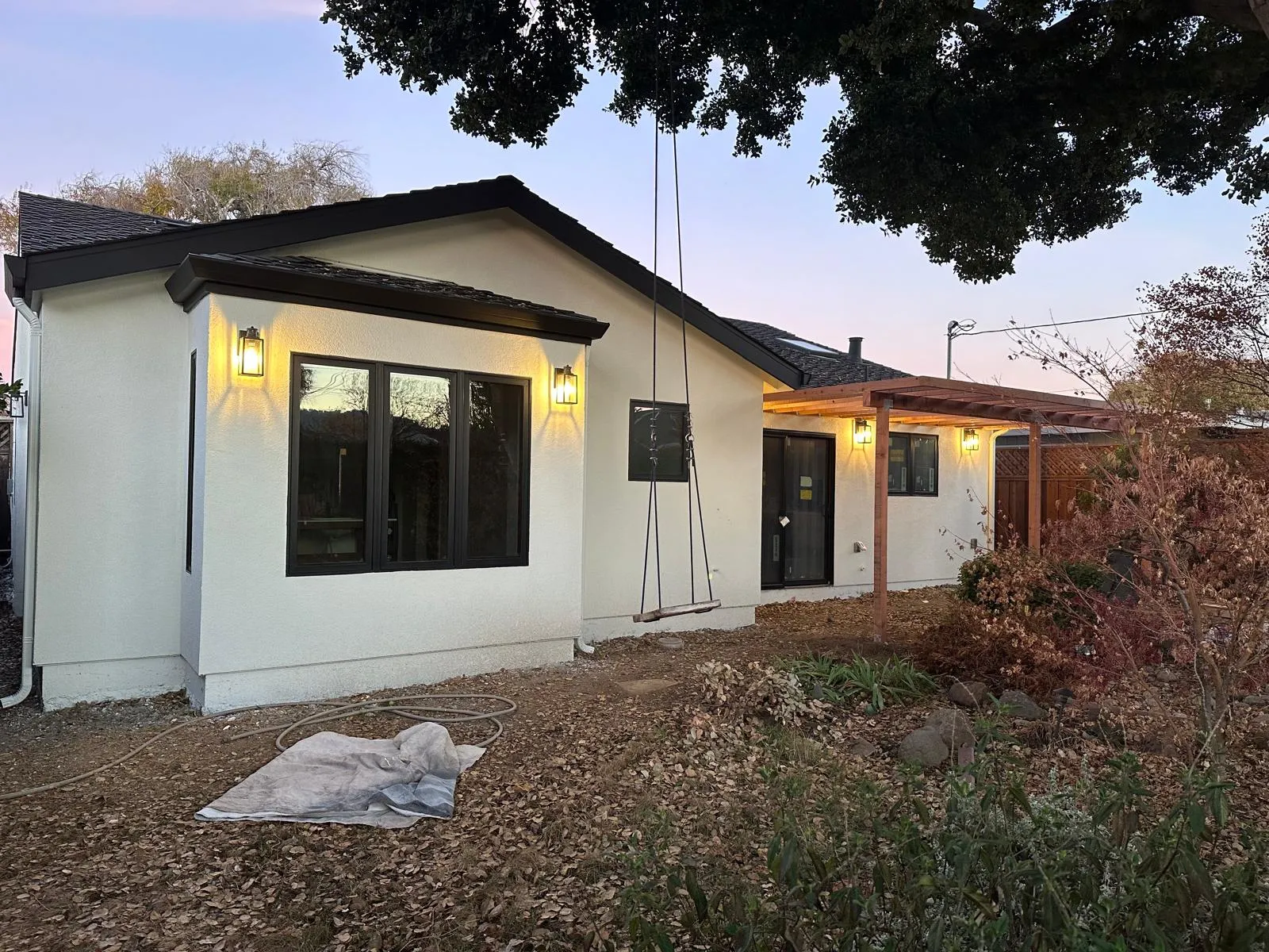 Rear exterior at dusk of a single-storey home after a remodel and addition — new white stucco, black trim and gutters, black-framed windows and slider, and a redwood pergola with lit sconces.