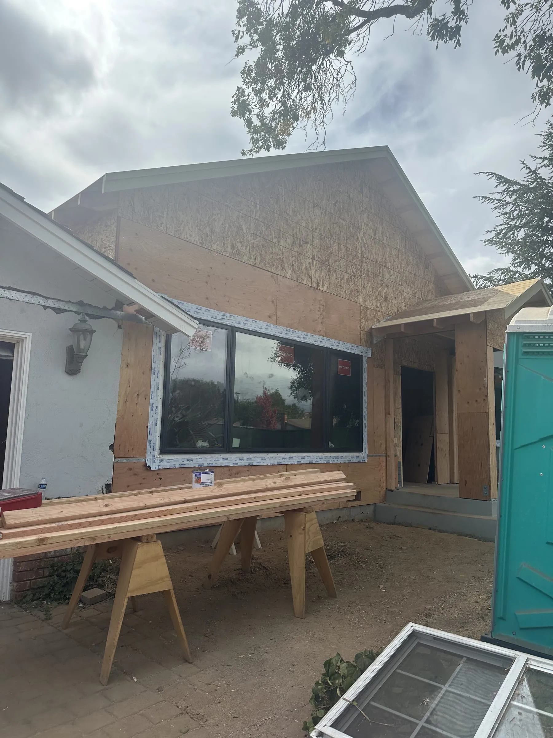 An addition mid-build: a new black-framed picture window set and taped into plywood sheathing under a newly framed gable, a covered entry porch framed alongside it, and lengths of trim stock laid across sawhorses in the yard.