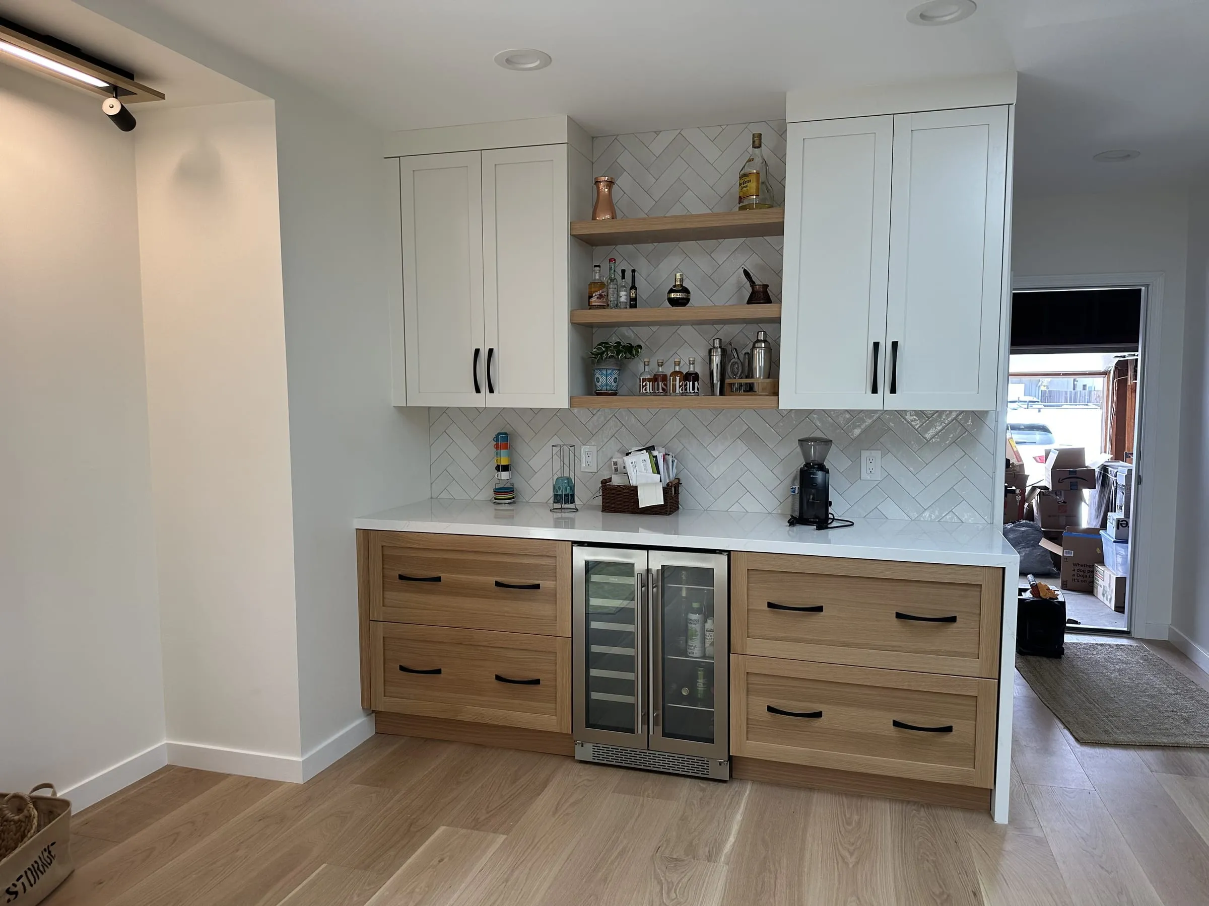 Finished home bar with white shaker upper cabinets, white oak drawer bases, a herringbone marble tile backsplash carried up between the uppers, three floating white oak shelves and an undercounter glass-front wine fridge.
