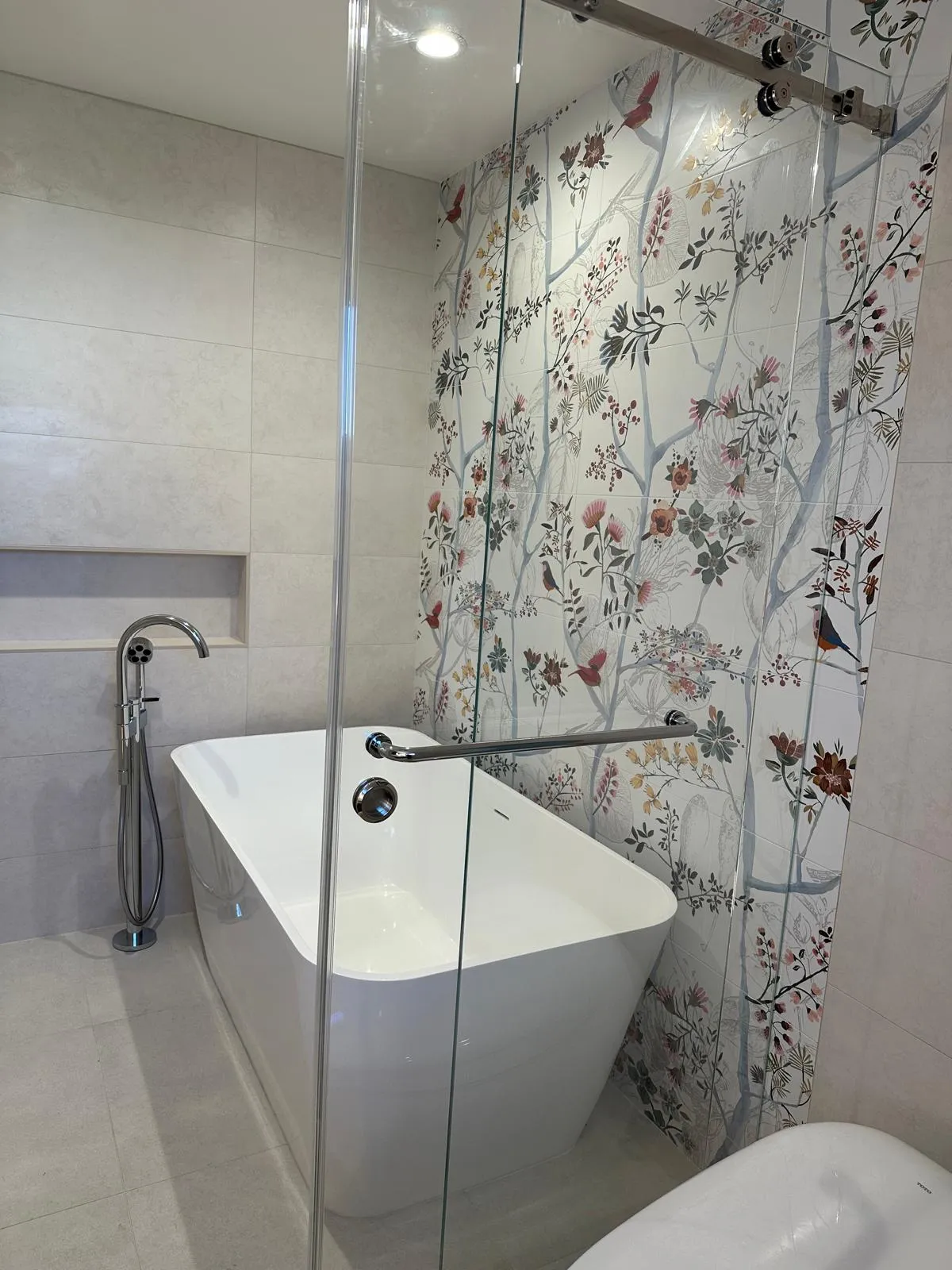 Completed bathroom with a freestanding tub, chrome floor-mounted tub filler, large-format cream tile and a botanical bird-and-branch feature tile wall behind frameless sliding glass.