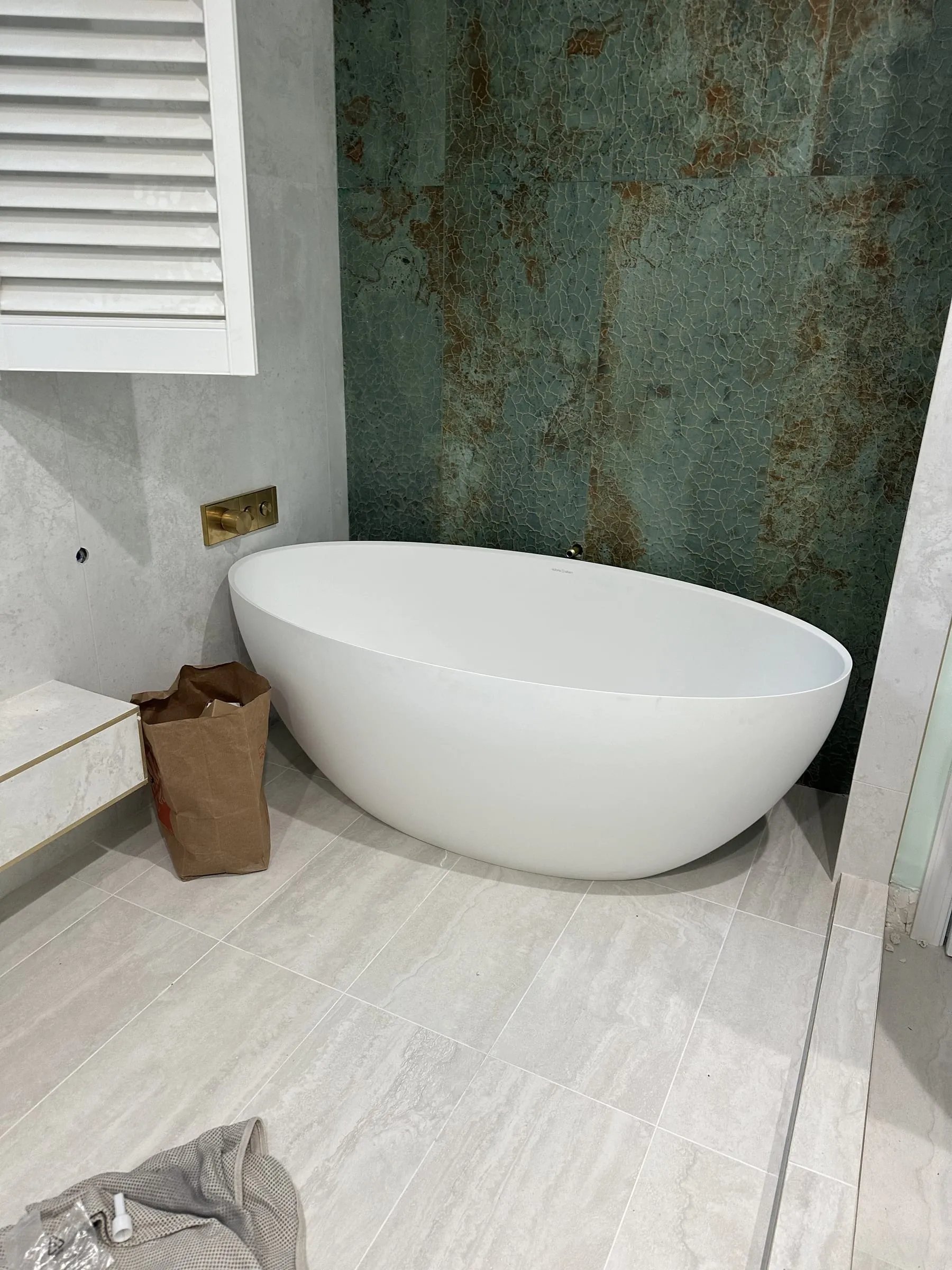 Primary bathroom with a matte white freestanding soaking tub set against a full-height green quartzite feature wall, travertine-look large-format tile on the surrounding walls and floor, a brass wall-mounted filler valve and white plantation shutters.