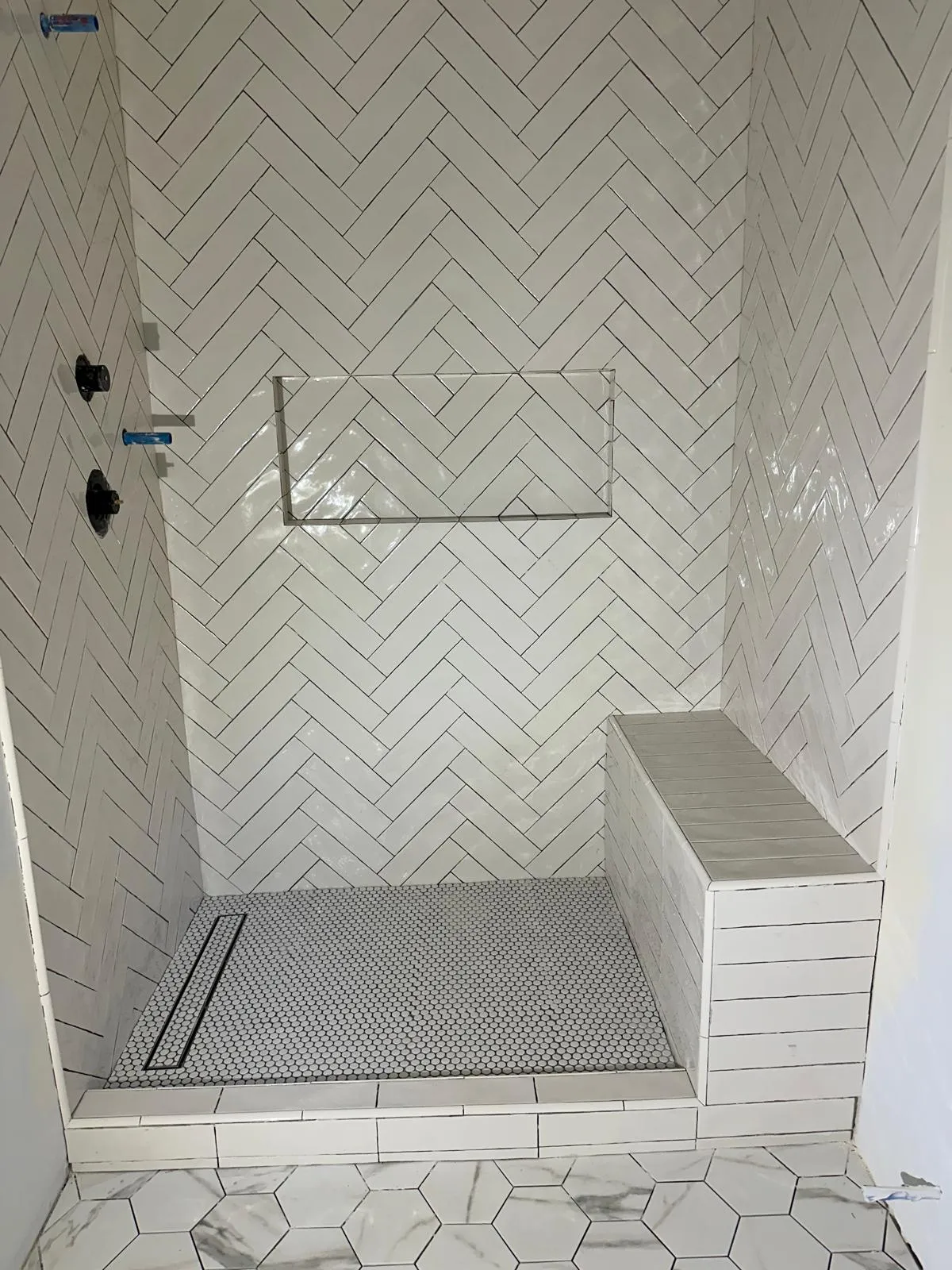 Shower enclosure under construction with white herringbone wall tile, a recessed niche, a tiled bench, grey penny-round floor tile with a linear drain and marble hex tile outside the curb.