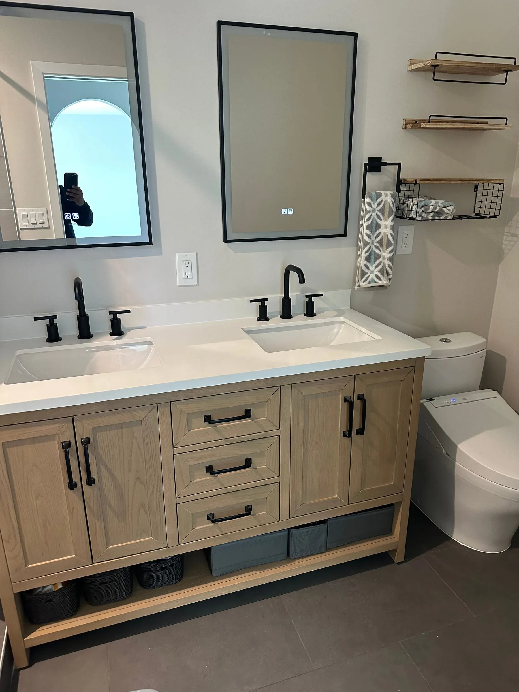 Finished bathroom with a white oak double vanity and quartz top, two undermount sinks with matte black faucets, a backlit LED mirror beside a black-framed mirror, oak shelves on black brackets and large-format grey floor tile.