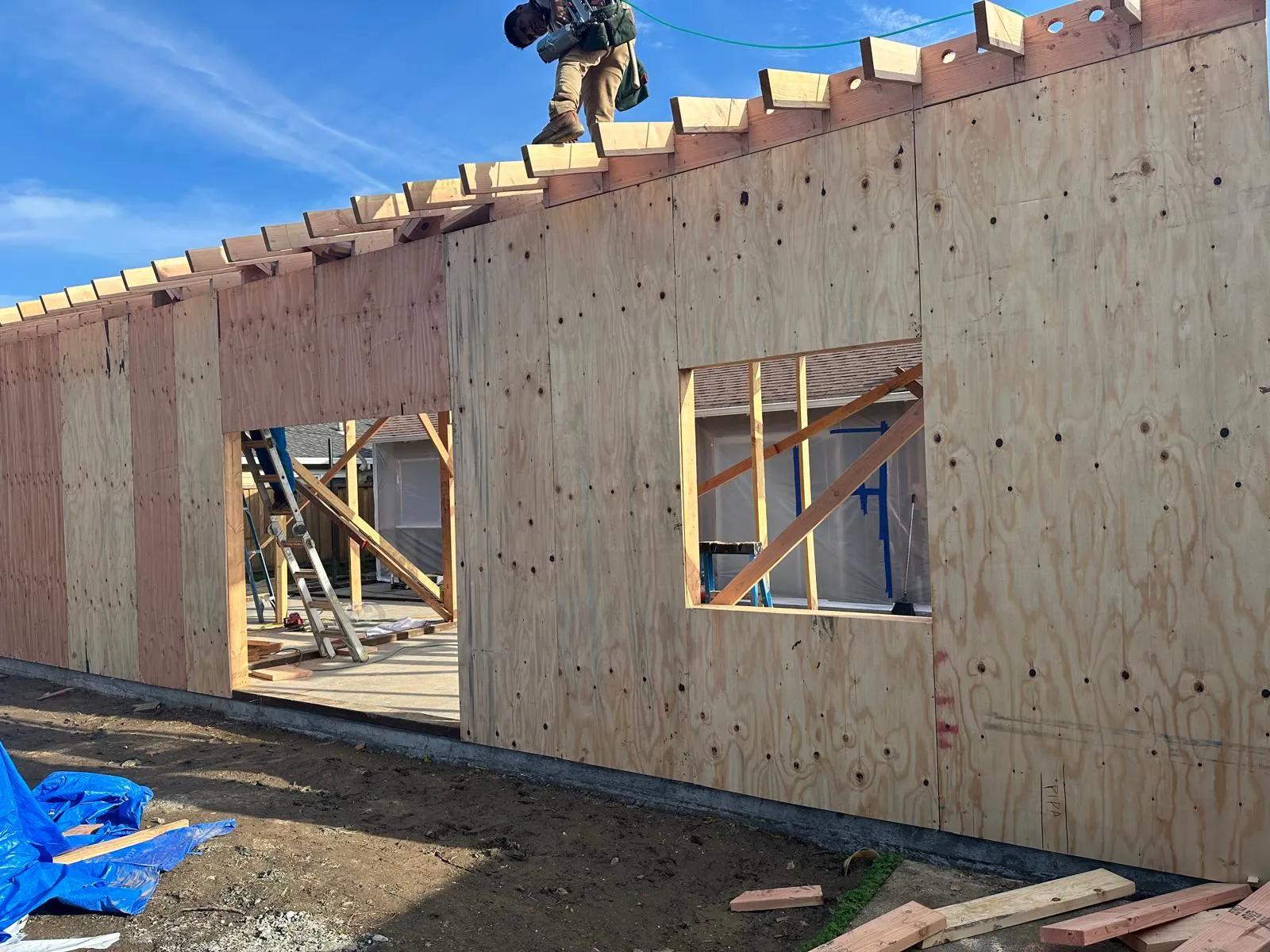 New structure being framed — plywood shear walls sheathed and nailed, roof rafters and outlookers set, and a framer walking the top plate with a nail gun.