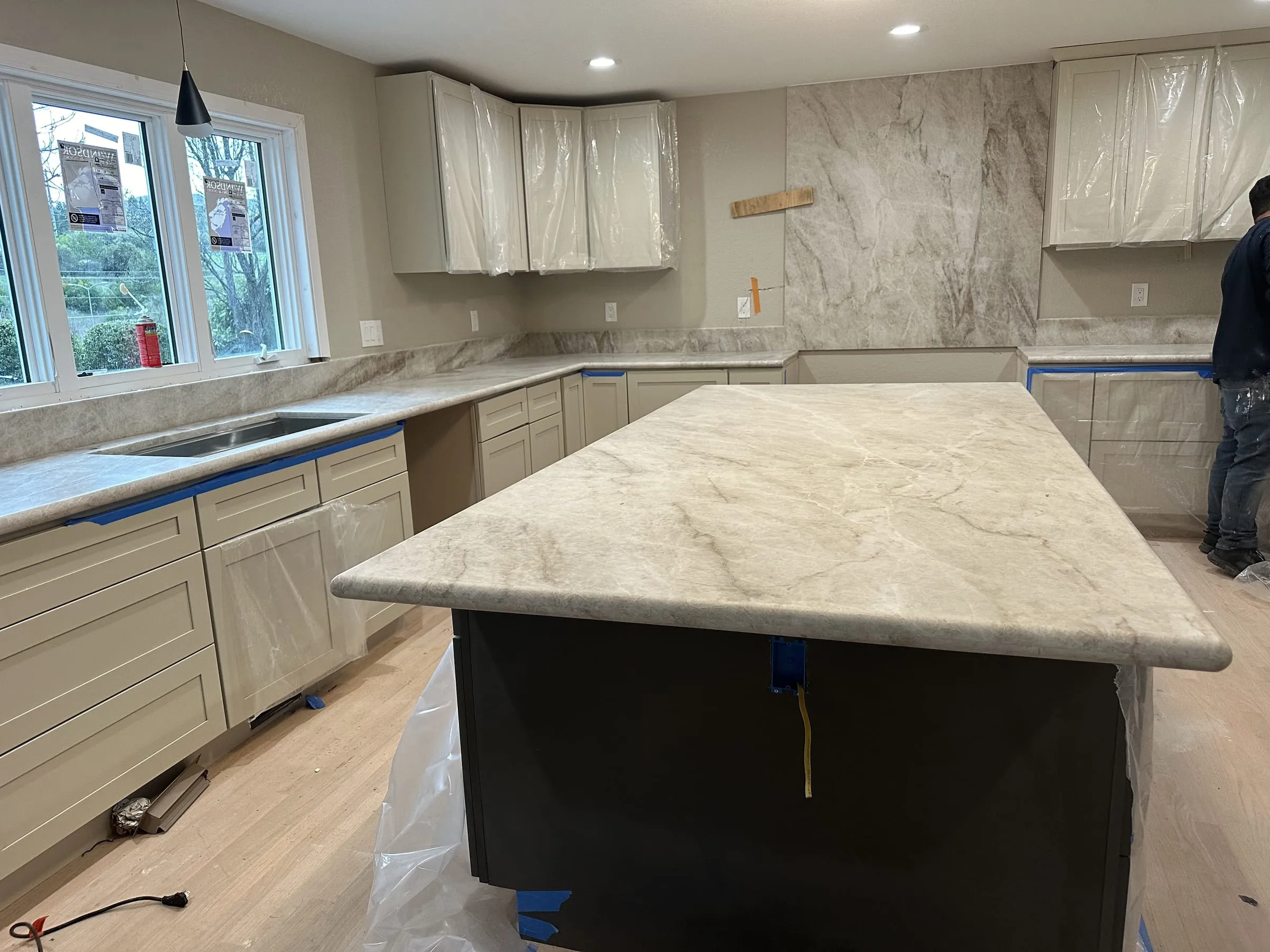 Kitchen under construction with a thick quartzite island top newly set on a black island base, matching stone counters along two walls, a full-height stone backsplash panel behind the range wall, and cabinets still masked in plastic sheeting.