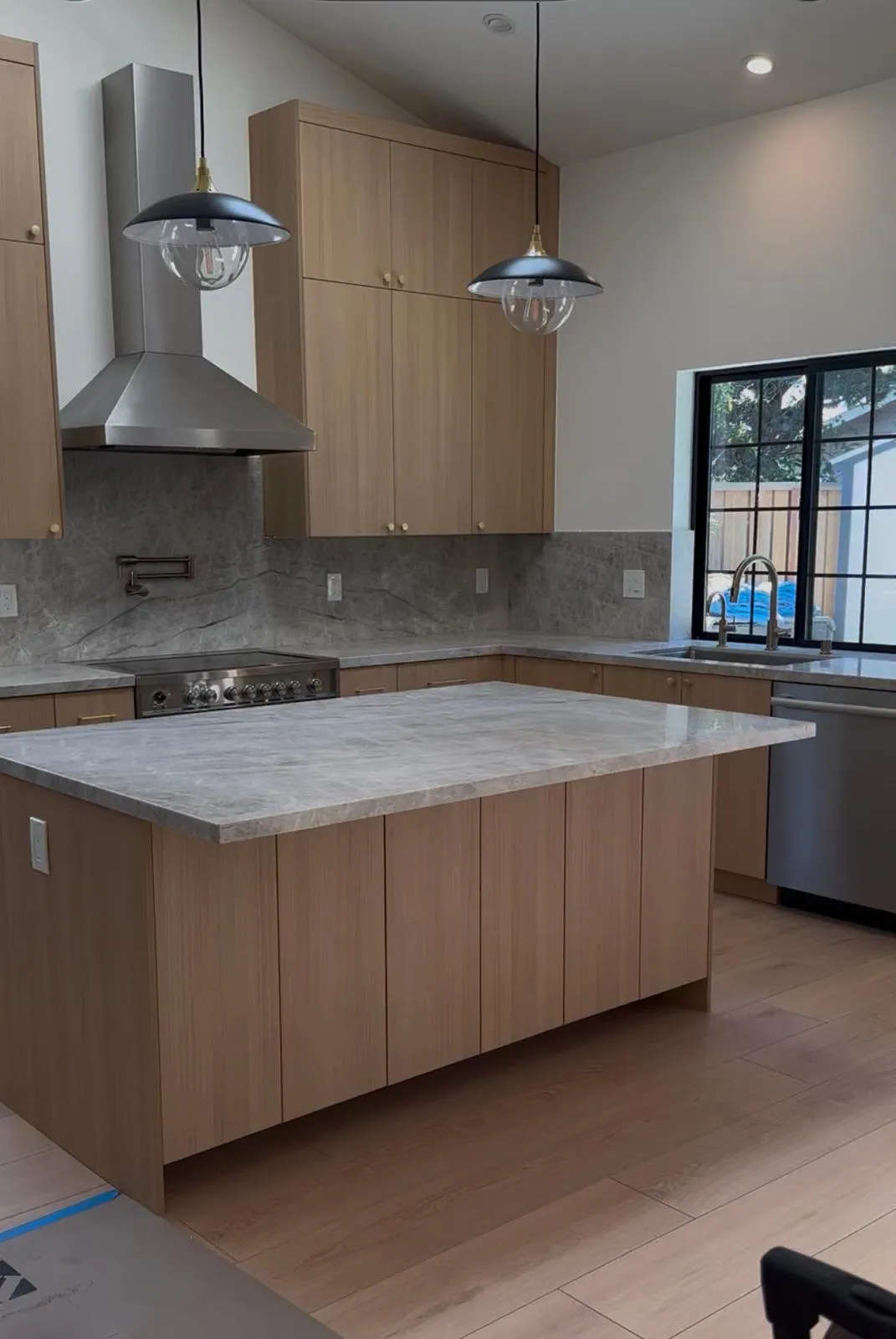 Finished kitchen with rift white oak flat-panel cabinetry, grey quartzite counters and full-height backsplash, stainless range and chimney hood, and a black steel-framed window over the sink.
