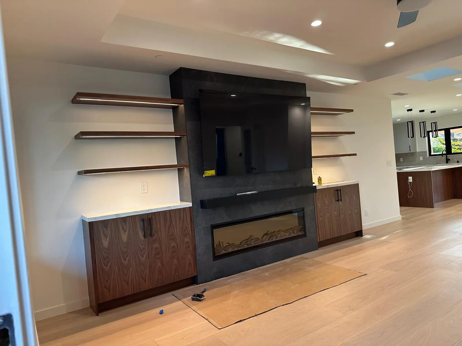 Great-room media wall with a black porcelain slab surround, linear electric fireplace, LED-lit walnut floating shelves and flanking walnut cabinets with stone tops.