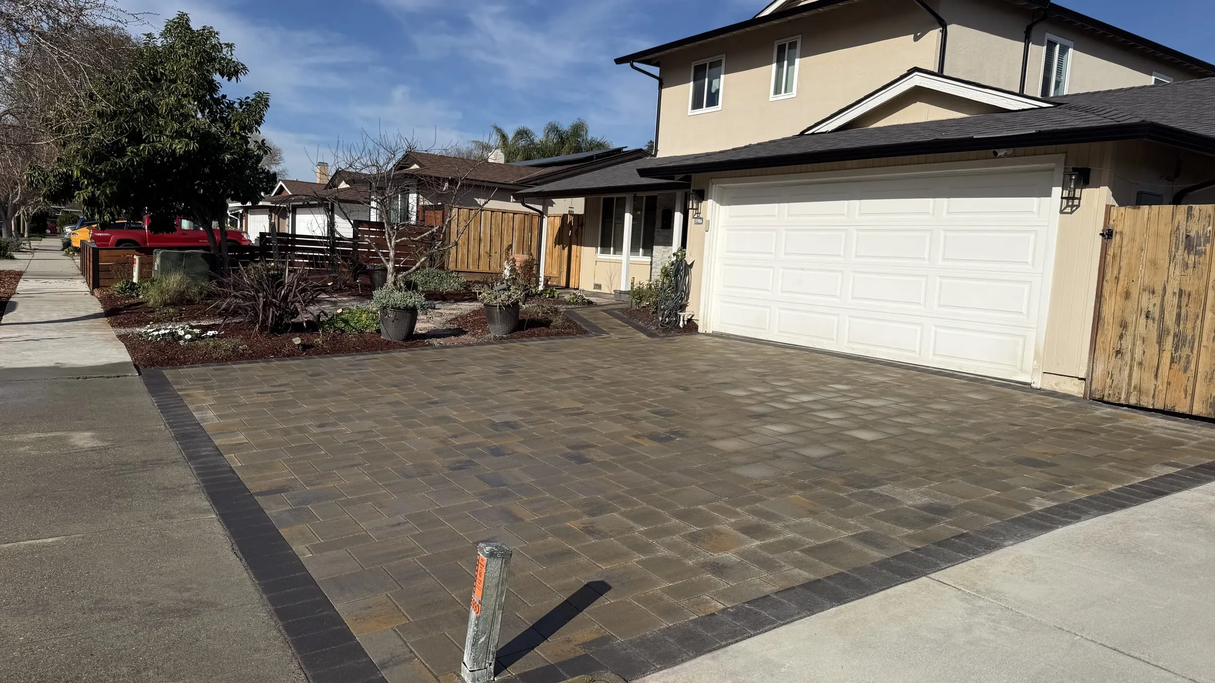 Finished driveway in tumbled tan and grey concrete pavers laid in a random ashlar pattern with a charcoal soldier-course border, running from the street to a two-car garage, with replanted beds and fresh bark mulch along the front of the house.